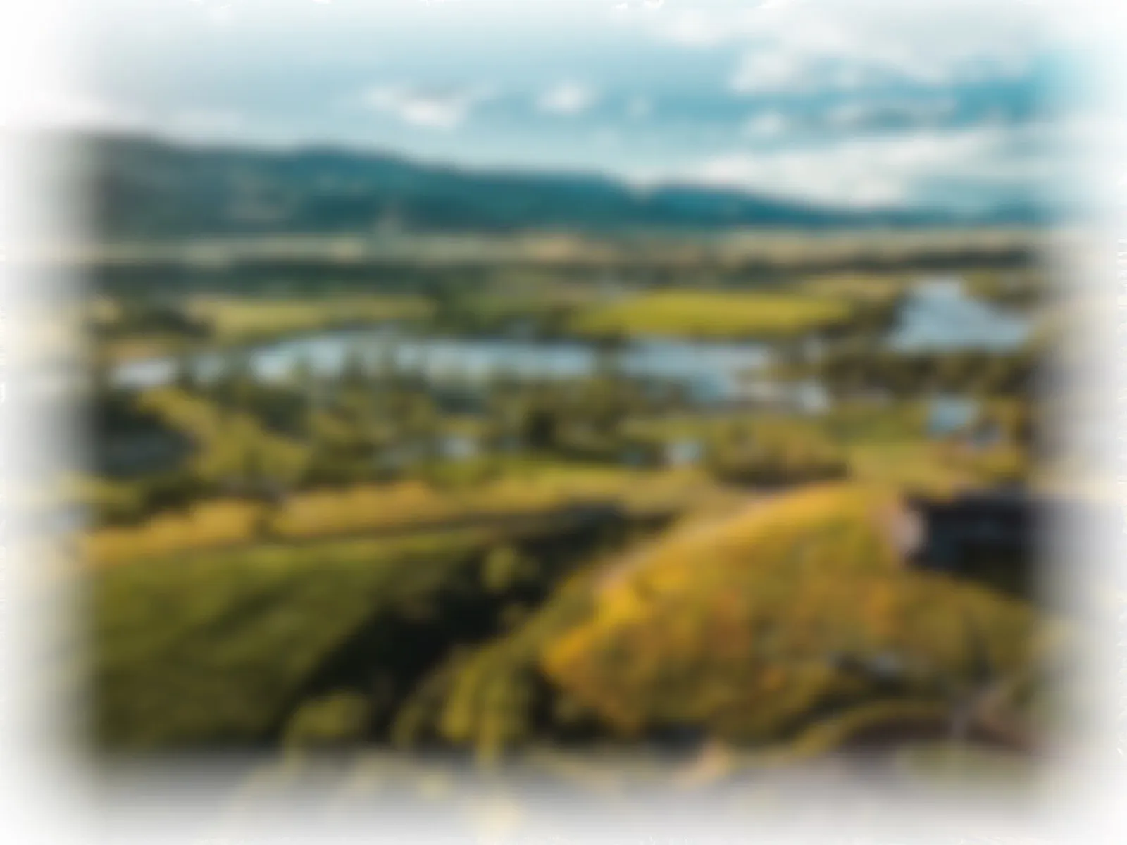 Blurry scenic view of a race course with water features and rolling hills under a cloudy sky.