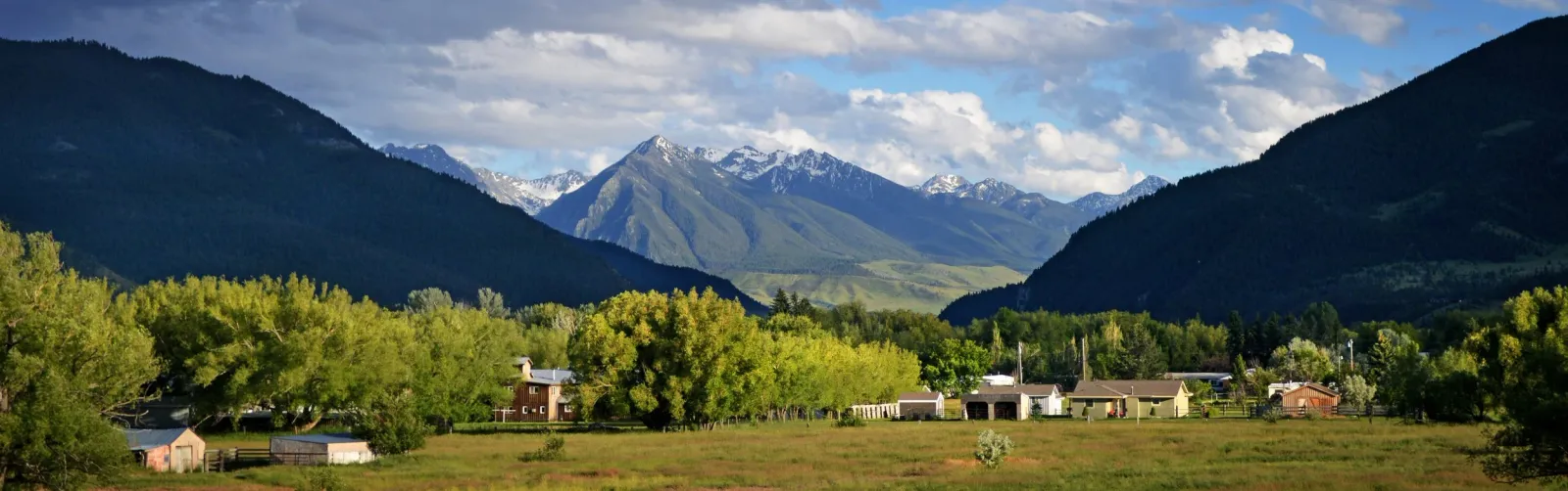 Panoramic view of a valley with trees and buildings, leading to snow-capped mountains under a cloudy sky.