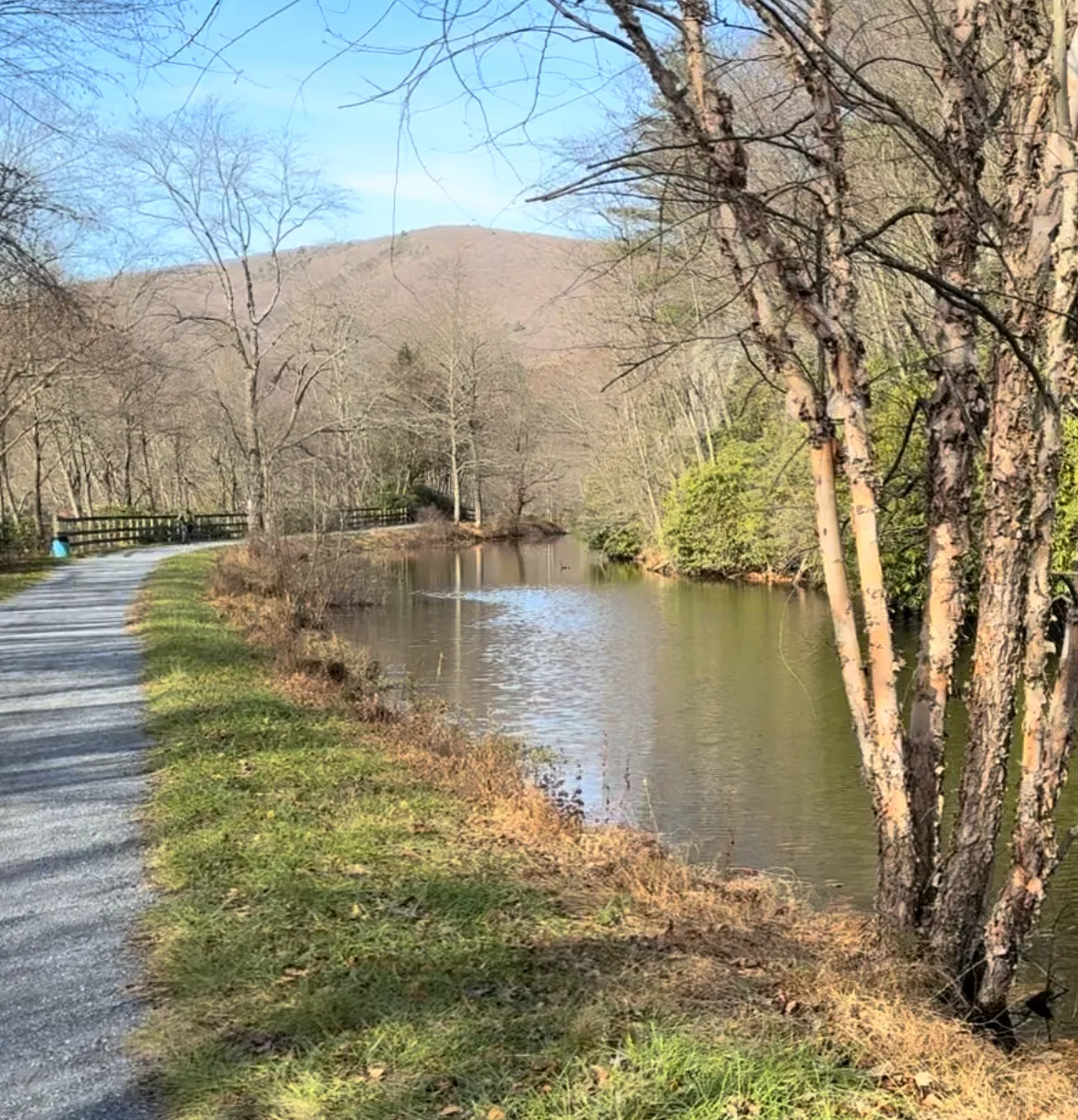 A gravel path runs alongside a calm body of water, with trees and a distant hill under a clear blue sky.