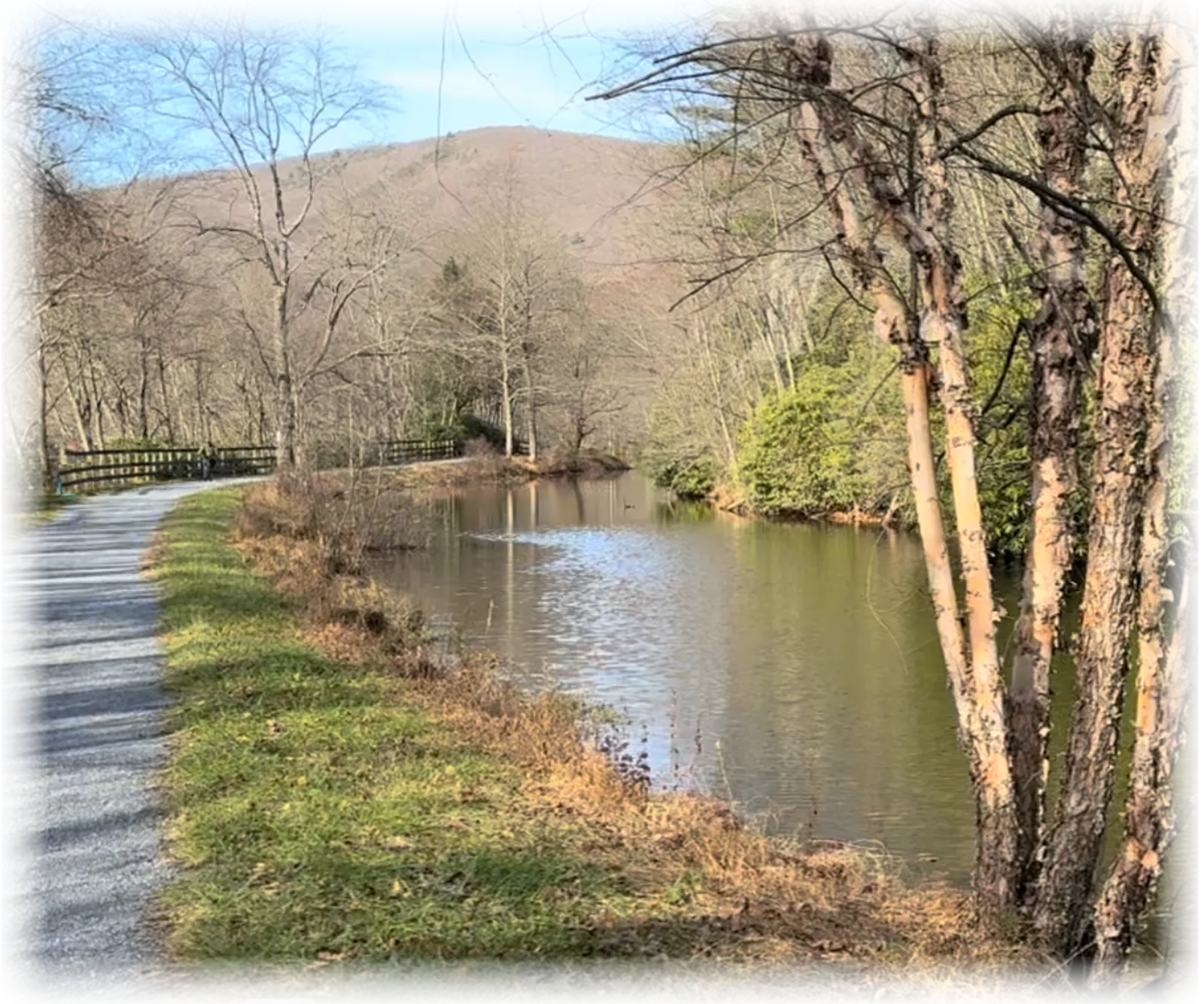A paved path runs alongside a calm body of water, with trees and a distant hill in the background.