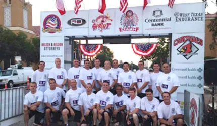A group of people in matching t-shirts stand in front of a race archway with various sponsor logos and flags.