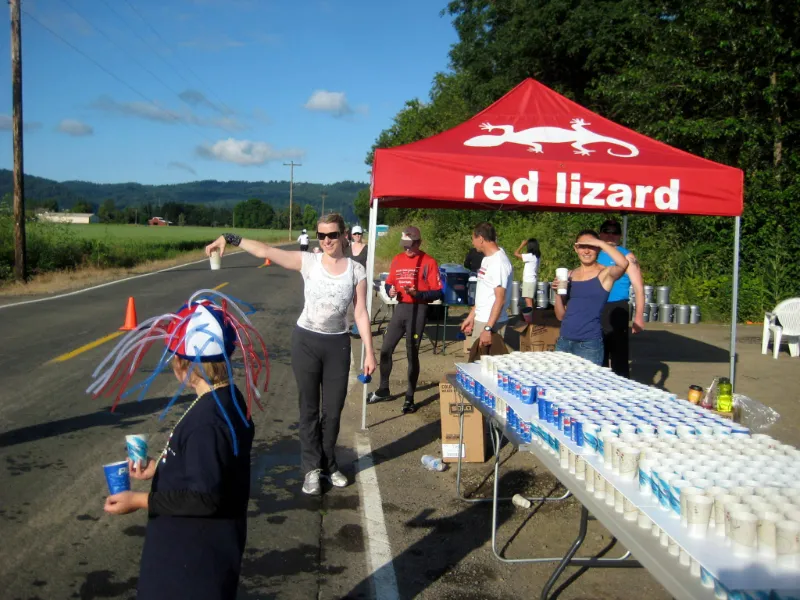 Runners and volunteers at an aid station during a road race, with a red sponsor tent and tables of cups.