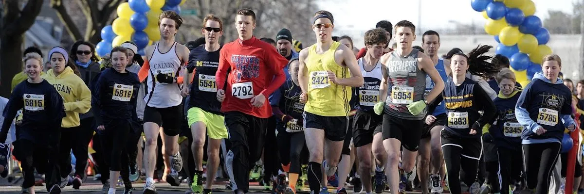 A group of runners, including men, women, and children, start a race with yellow and blue balloons overhead.