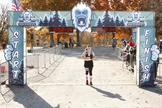 A female runner crosses the finish line at the Yeti Endurance Weekend race, with the start/finish archway and timers visible.