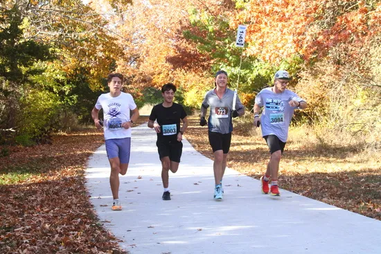 Four male runners are running on a paved path lined with trees in autumn colors.