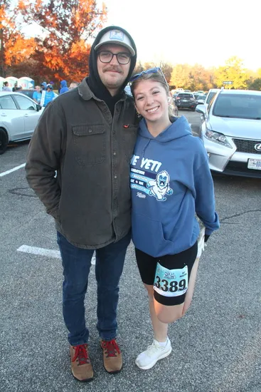 A smiling couple poses for a photo, the woman on the right wears a blue hoodie with 'YETI' logo and a race bib number 3389, stretching her leg.
