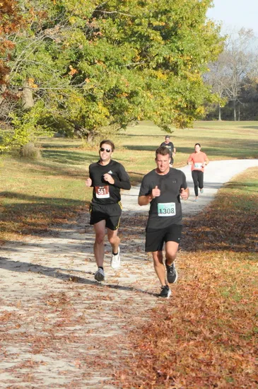 Two male runners are running on a dirt path lined with autumn leaves, with other runners in the background.