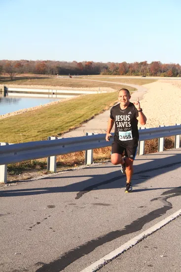 A runner wearing a black t-shirt with 'JESUS SAVES' printed on it, running on a paved road with a body of water and trees in the background.