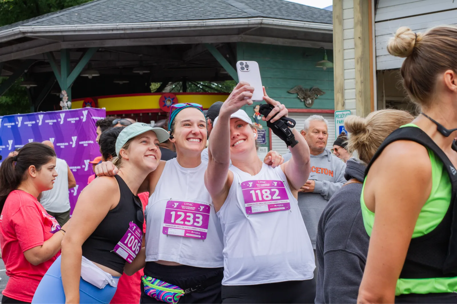 Three women smiling and taking a selfie at the start line of a YMCA Harrisburg race, wearing bib numbers.