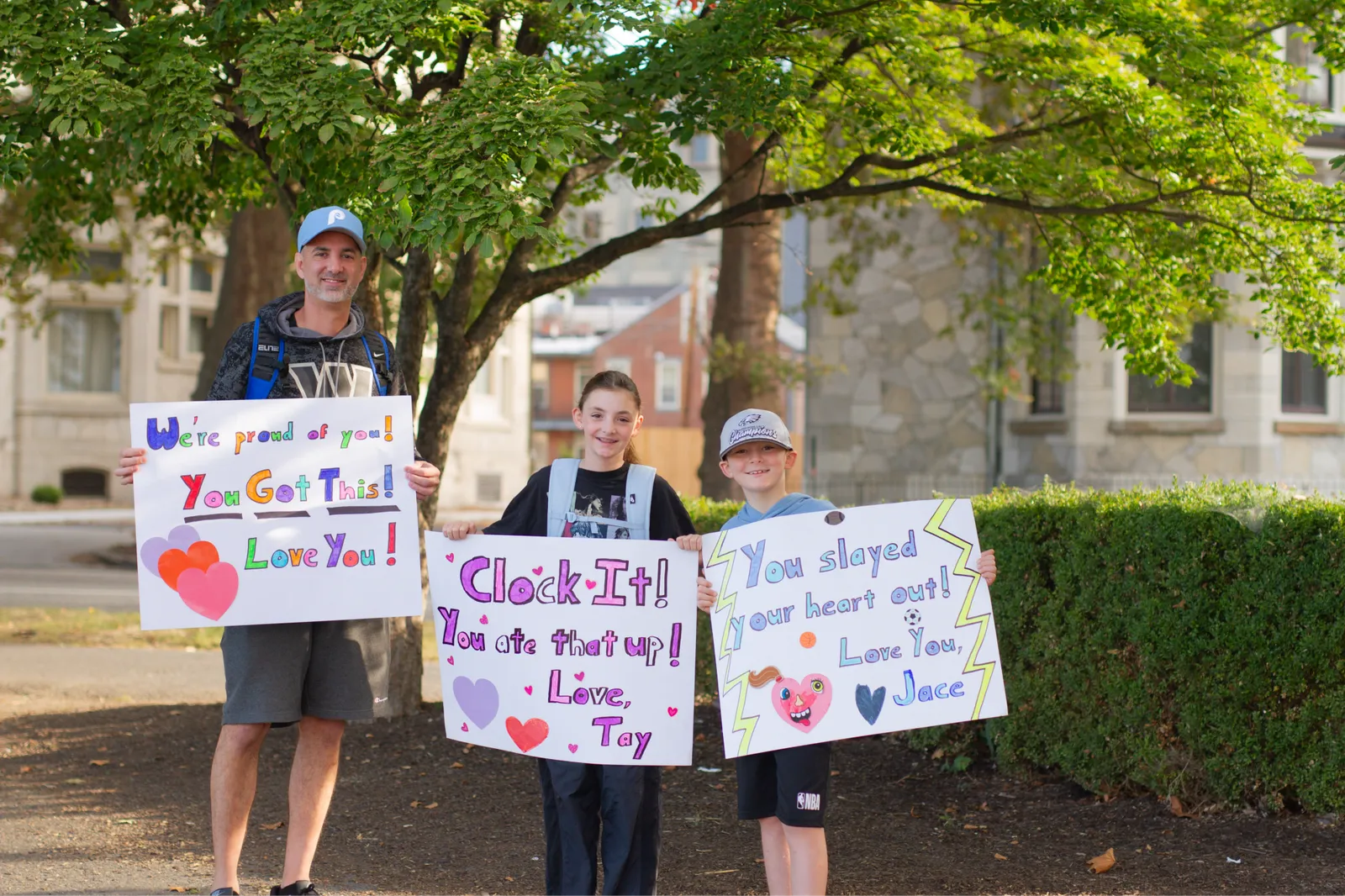 A man and two children hold up colorful signs with encouraging messages for runners, such as 'We're proud of you!' and 'You Got This!'