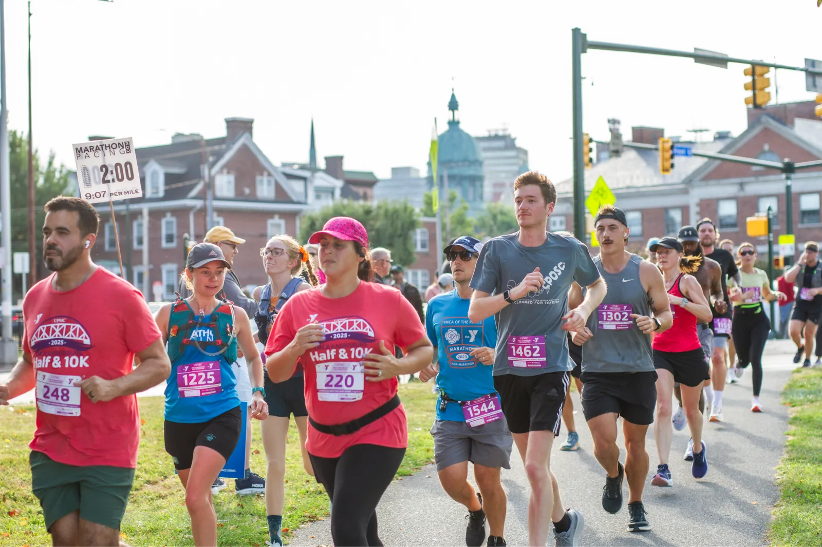 A group of runners participating in a half marathon or 10k race, with some wearing race bibs and others looking ahead as they run on a paved path.