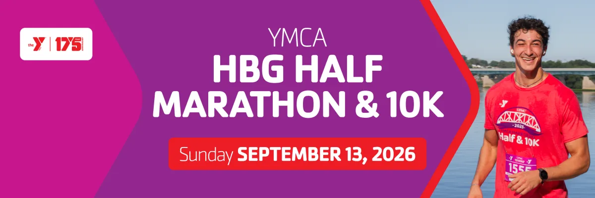 A smiling young man wearing a red YMCA Half & 10K race shirt and bib number stands by the water, looking towards the camera. The race name and date are visible on a purple banner in the background.