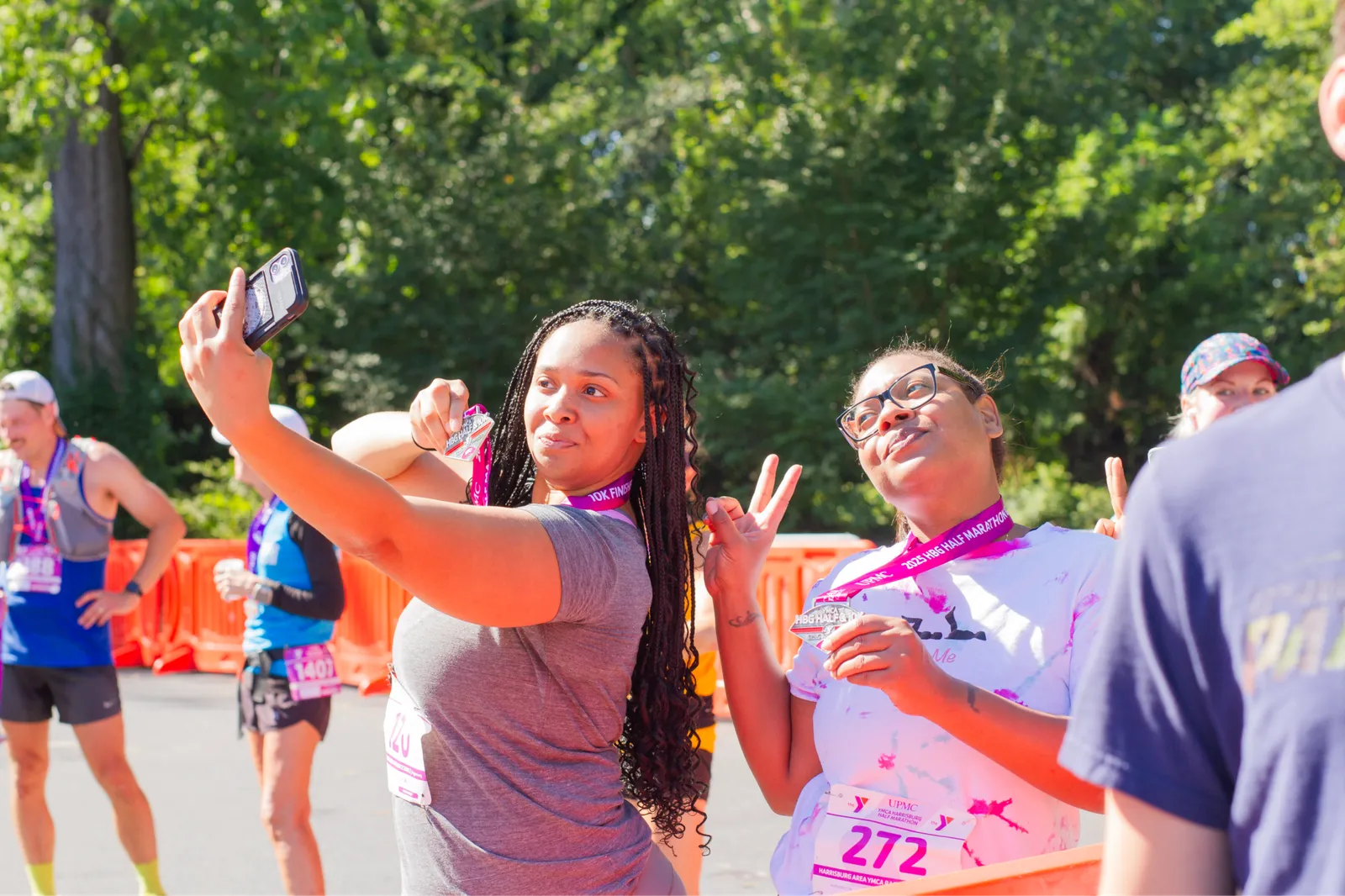 Two women smiling and holding up their race medals, one taking a selfie with her phone, with other runners in the background.