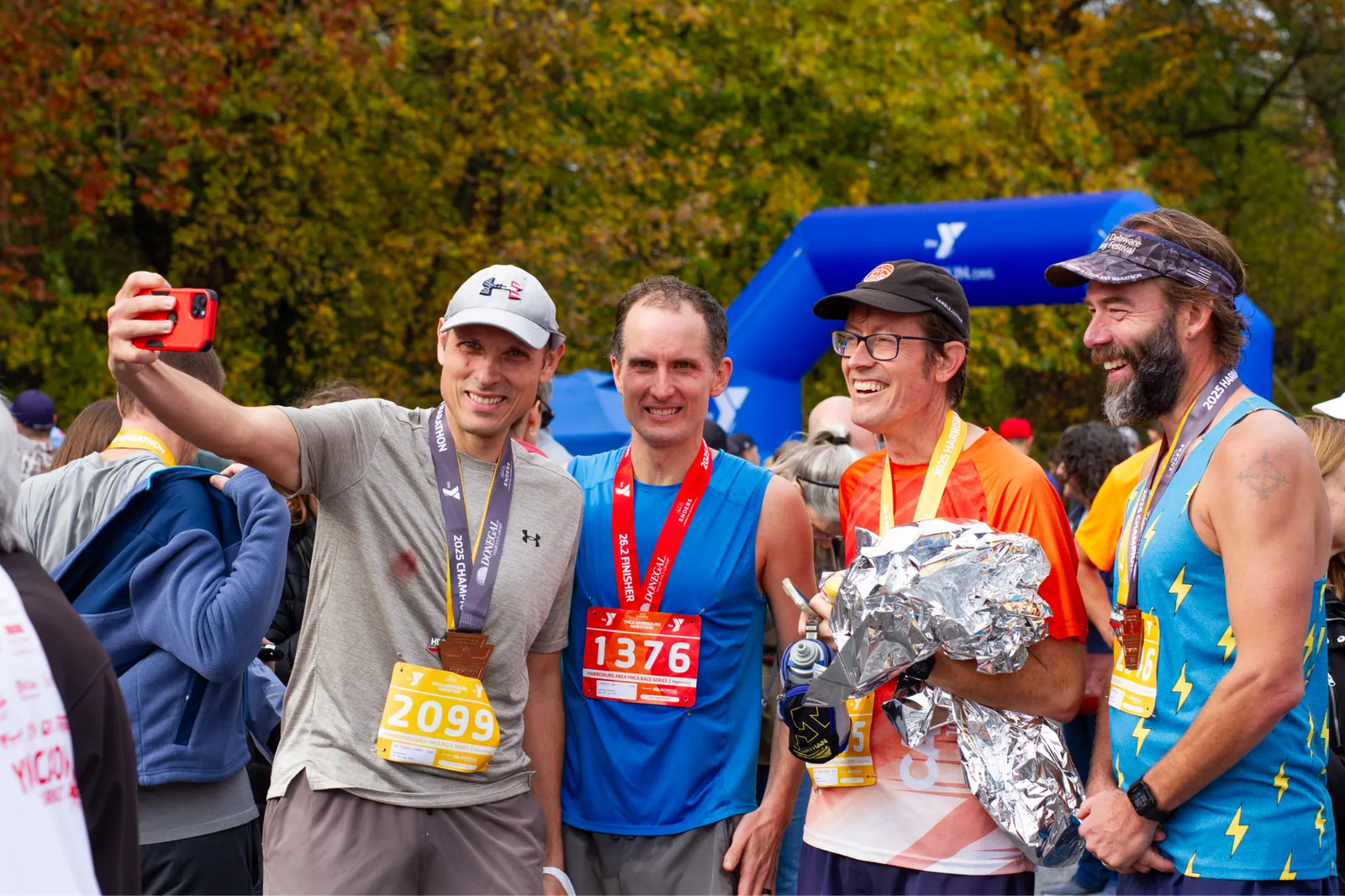Four runners smiling and posing for a selfie after completing a race, with medals around their necks and a YMCA arch in the background.