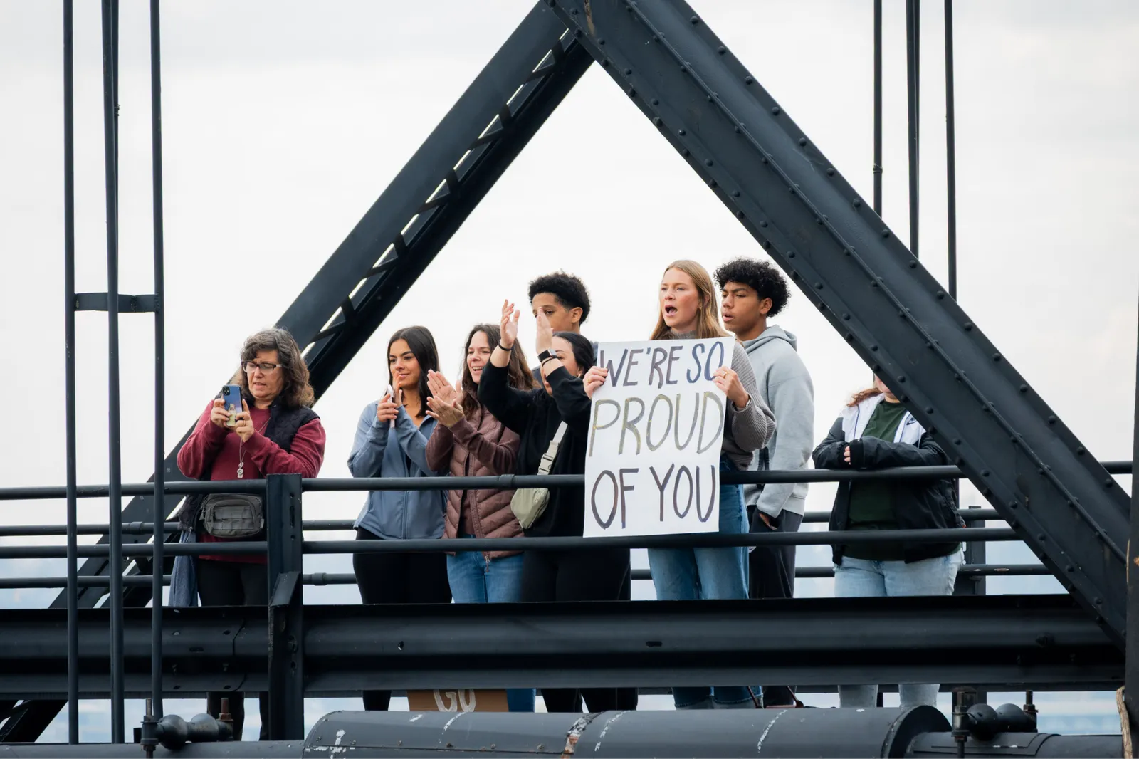 A group of people on a bridge, some holding a sign that says 'WE'RE SO PROUD OF YOU', cheering for runners.