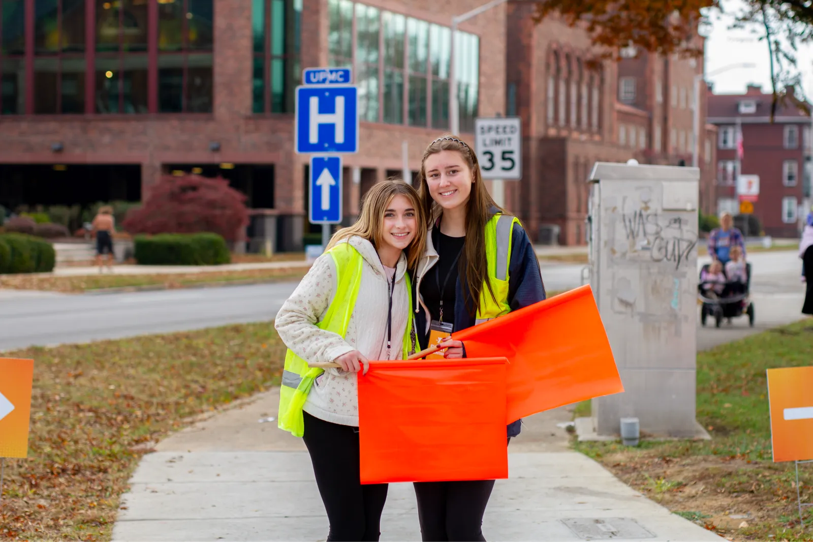 Two young women in safety vests smiling and holding orange flags on a sidewalk with an urban background.