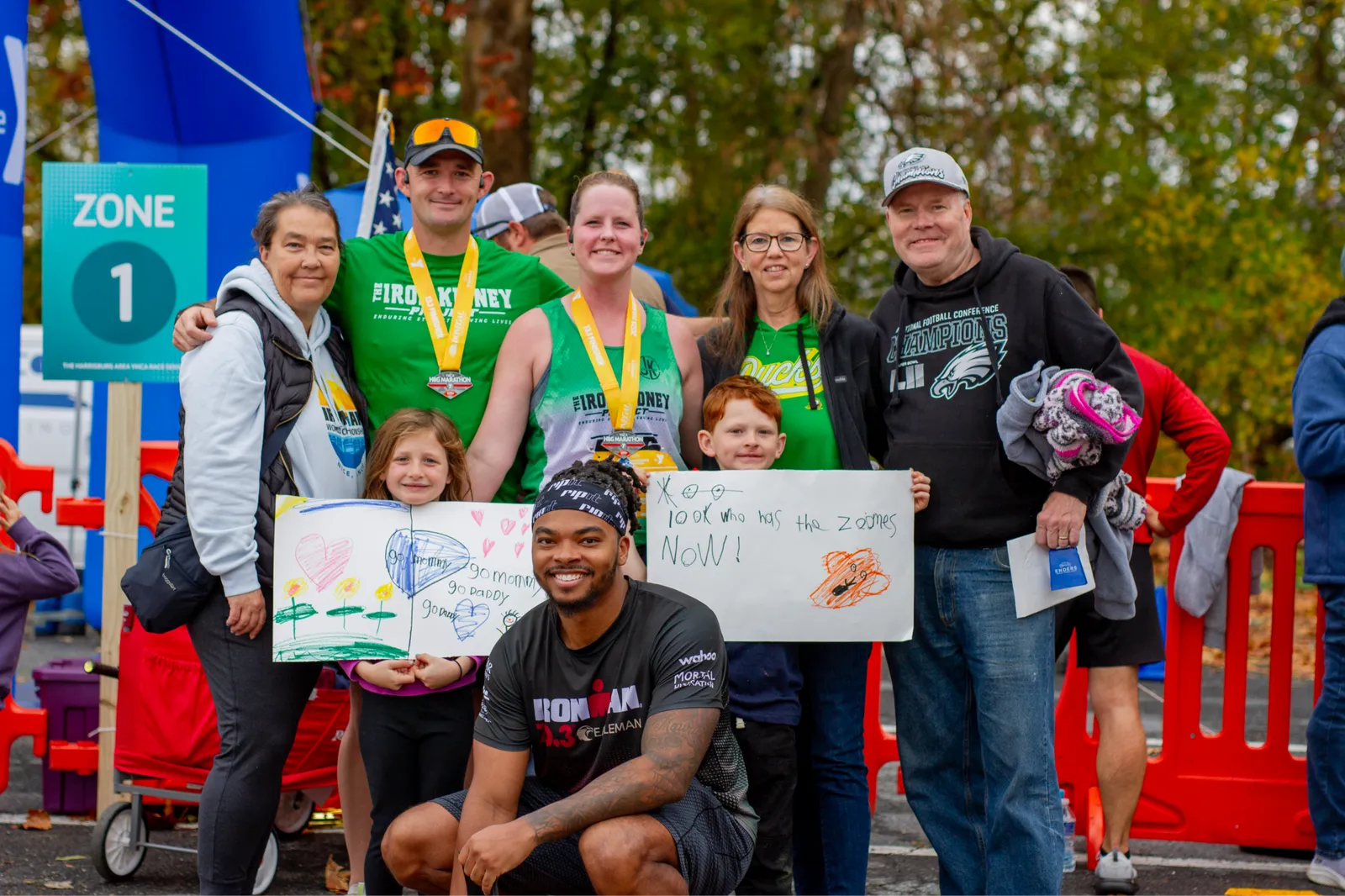A group of runners and their families pose for a photo after a race, holding up handmade signs and wearing medals.