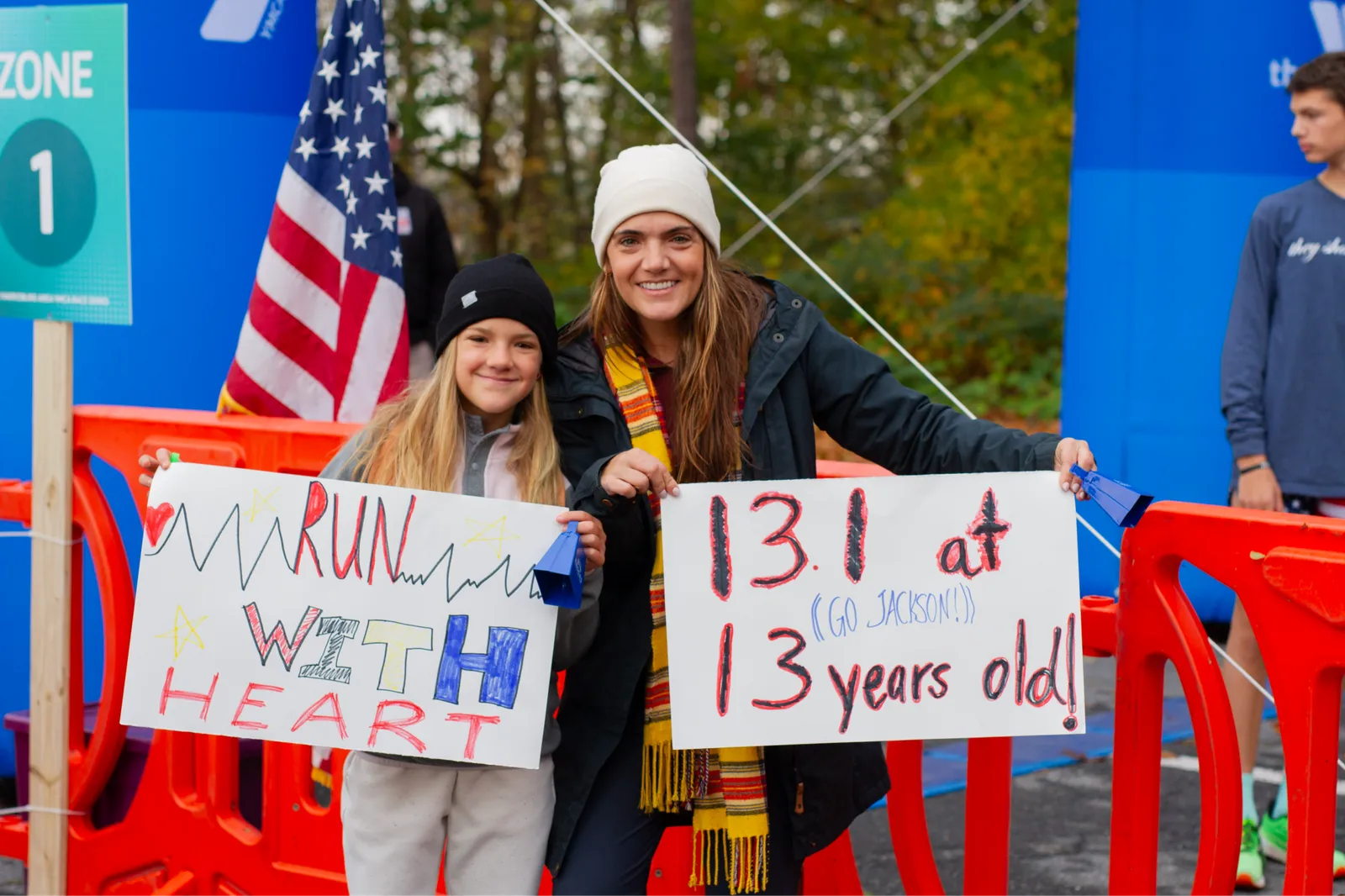 A woman and a young girl smile while holding signs at a race event. The girl's sign says "RUN WITH HEART" and the woman's sign says "13.1 at 13 years old! (GO JACKSON!)".