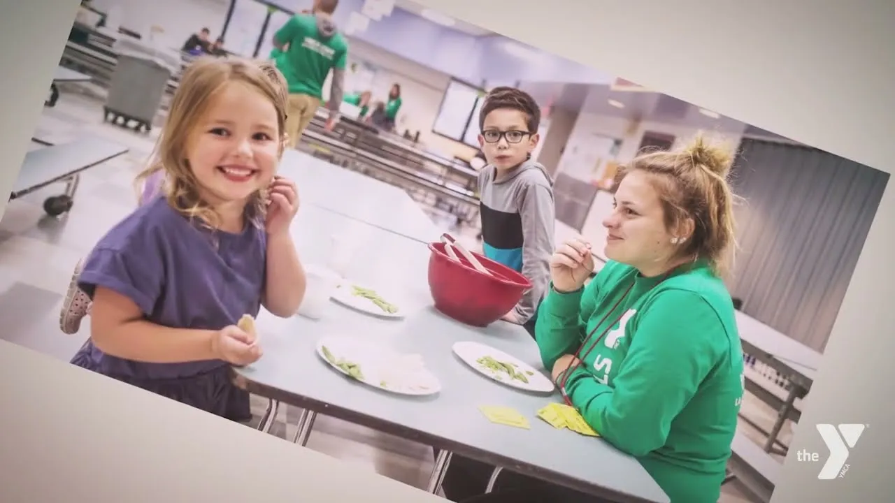 A young girl smiles at the camera while sitting at a table with other children and an adult.
