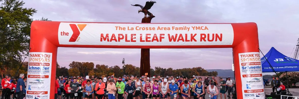 A large group of runners gathered at the start line of the YMCA Maple Leaf Walk Run, beneath a red inflatable archway with the race name and sponsor logos.