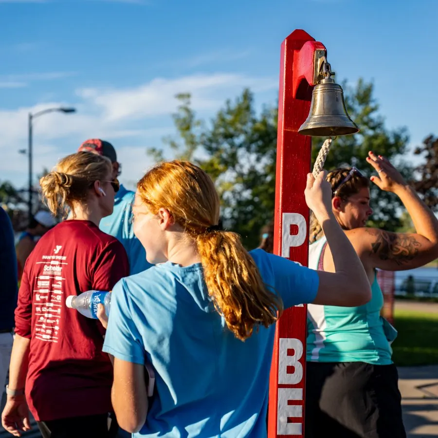 People gathered around a red post with a bell, with one person pulling the rope, suggesting a pre-race ceremony or start-line event.