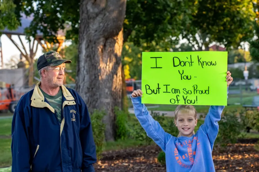 A young girl holds up a bright green sign that says 'I Don't Know You - But I Am So Proud of You!' while an adult man stands beside her, both smiling.
