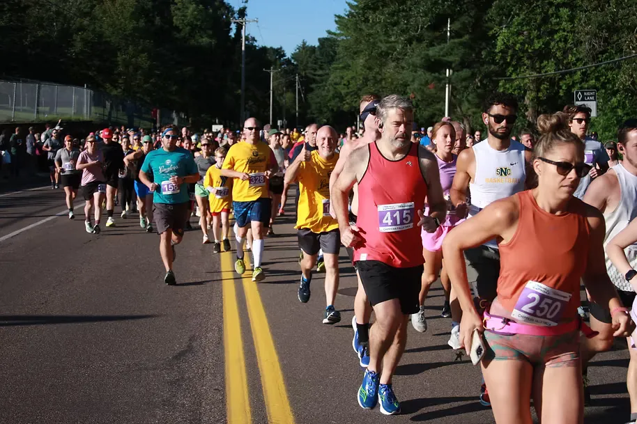 A group of runners participating in a road race, with some runners in the foreground and a larger crowd in the background.