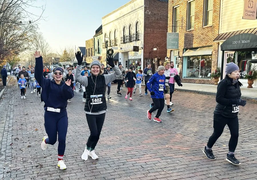 Runners smiling and waving as they participate in a race on a brick-paved street lined with shops.