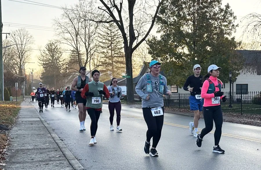 A group of runners on a paved road during a race, with trees and houses in the background.